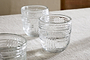 A set of four nkuku Kaanch Tumblers in clear glass, featuring an embossed check pattern and thick bases, sit on neutral fabric. Their textured surfaces reflect light beautifully, creating striking highlights and shadows.