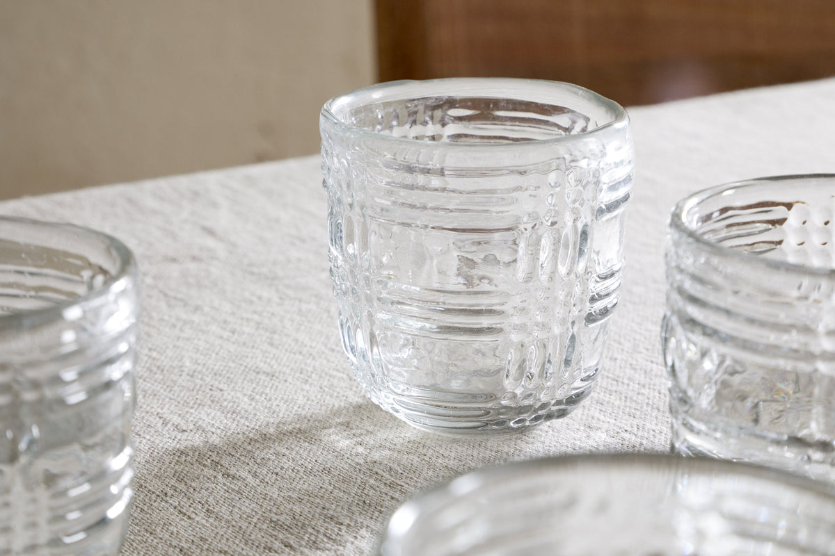 The nkuku Kaanch Tumbler - Clear (Set of 4) features embossed check pattern glassware with a thick, sturdy design, displayed on a woven tablecloth with natural light highlighting their craftsmanship and subtle wooden accents in the background.
