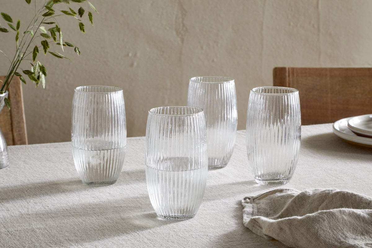 A set of four nkuku Otuba Highball Clear glasses is arranged on a light beige tablecloth, each half-filled with water. Wooden chairs, stacked plates, a cloth napkin, and a small vase of greenery complete the elegant table setting.