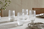 A set of four nkuku Otuba Highball Clear glasses is arranged on a light beige tablecloth, each half-filled with water. Wooden chairs, stacked plates, a cloth napkin, and a small vase of greenery complete the elegant table setting.