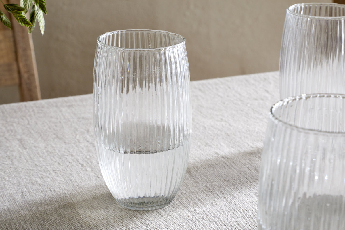 A nkuku Otuba Highball - Clear (Set of 4) glass sits half-filled with water on a light gray textured tablecloth, while three matching highball glasses are partially visible nearby, creating a calm, elegant atmosphere.