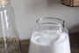 The nkuku Chaaval Storage Jar - Clear, filled with white granulated sugar, sits on a wooden surface. To the left, part of another jar with a brown coarse ingredient adds an organised touch to the kitchen scene.