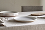 A neutral-toned dining table set with a light linen tablecloth, stacked ceramic plates, an nkuku Edo Dinner Plate in terracotta front and center, a wooden cutting board, silverware, and folded napkin highlights minimalist style. An upholstered chair sits behind.
