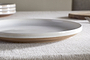 The nkuku Edo Dinner Plate - Terracotta features a white glazed ceramic surface with a brown unglazed rim, displayed on a textured beige tablecloth. In the background, stacked plates, a wooden board, and a fork appear softly out of focus.
