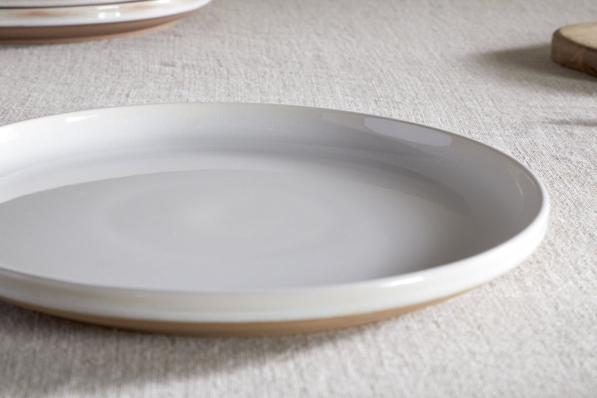 The nkuku Edo Dinner Plate - Terracotta features a minimalist round ceramic design with a glossy white surface and light brown rim, shown resting on a textured beige tablecloth with another plate and wooden board softly blurred in the background.
