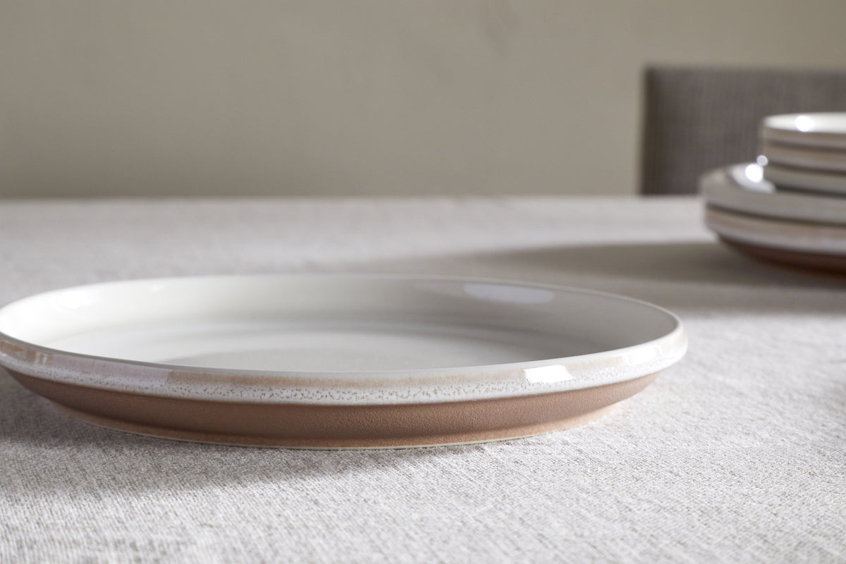 The nkuku Edo Side Plate - Terracotta features a white glazed stoneware interior and a brown unglazed rim, shown on a light textured tablecloth with stacked plates and a softly blurred beige chair enhancing the minimalist look.