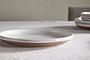 The nkuku Edo Side Plate - Terracotta features a white glazed stoneware interior and a brown unglazed rim, shown on a light textured tablecloth with stacked plates and a softly blurred beige chair enhancing the minimalist look.