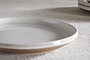 A close-up of the nkuku Edo Side Plate - Terracotta, featuring a beige and white glaze with a subtle brown rim and glossy finish, displayed on textured fabric. Another similar plate is partially visible in the background.