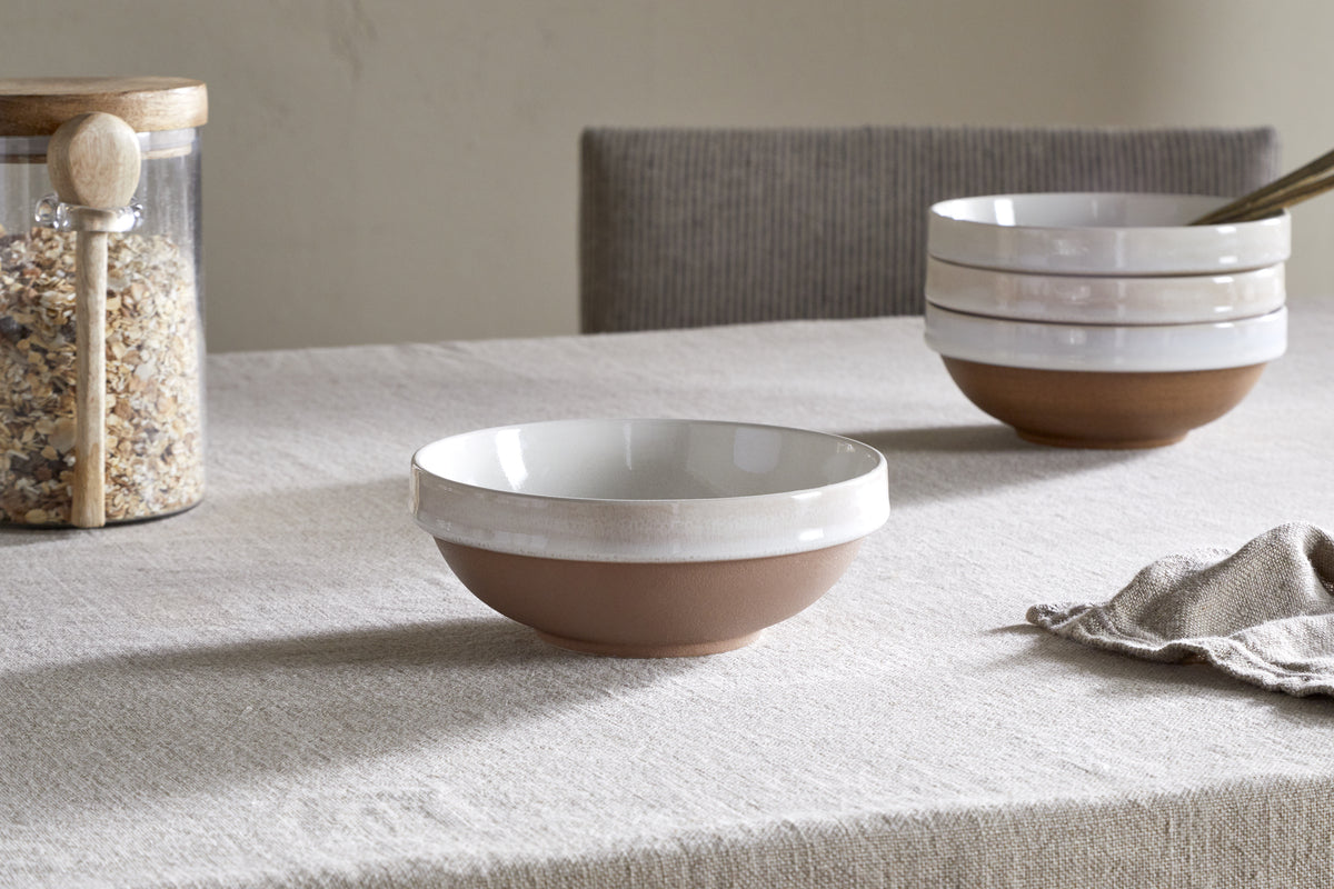 A beige tablecloth covers a table set with an nkuku Edo Cereal Bowl - Terracotta, showcasing its white glazed rim and brown base. Handcrafted bowls, a jar of oats with a wooden spoon, and a cloth napkin are seen in the background.
