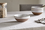 A beige tablecloth covers a table set with an nkuku Edo Cereal Bowl - Terracotta, showcasing its white glazed rim and brown base. Handcrafted bowls, a jar of oats with a wooden spoon, and a cloth napkin are seen in the background.
