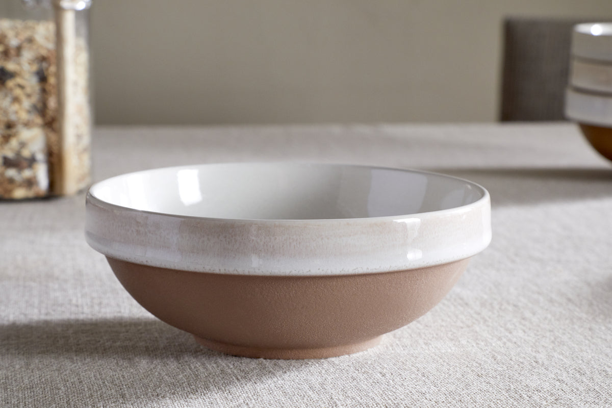 The nkuku Edo Cereal Bowl - Terracotta, featuring a matte brown base and glossy white top, sits empty and centered on a beige textured tablecloth with blurred plates in the background, showcasing minimalist tableware style.