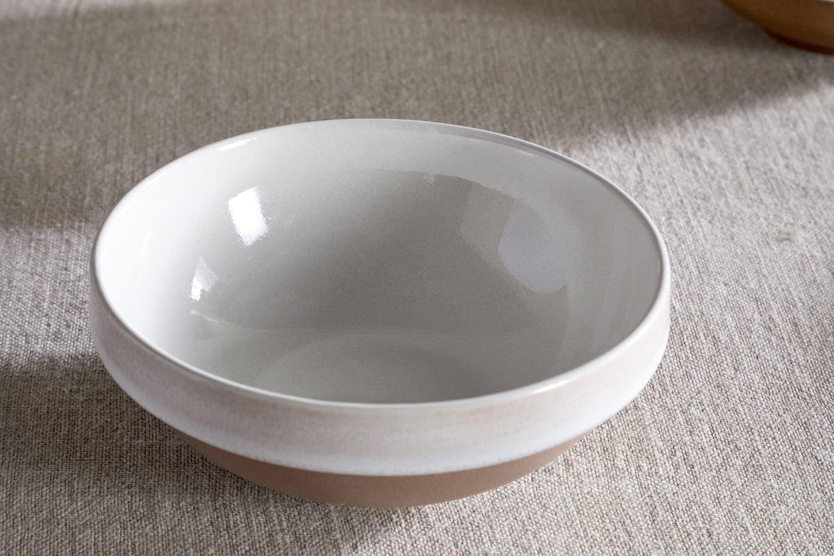 The nkuku Edo Cereal Bowl - Terracotta, featuring a glossy white interior and matte beige exterior, rests on a textured light fabric. Minimalist and empty, the bowl is gently lit by natural light, creating soft reflections on its surface.