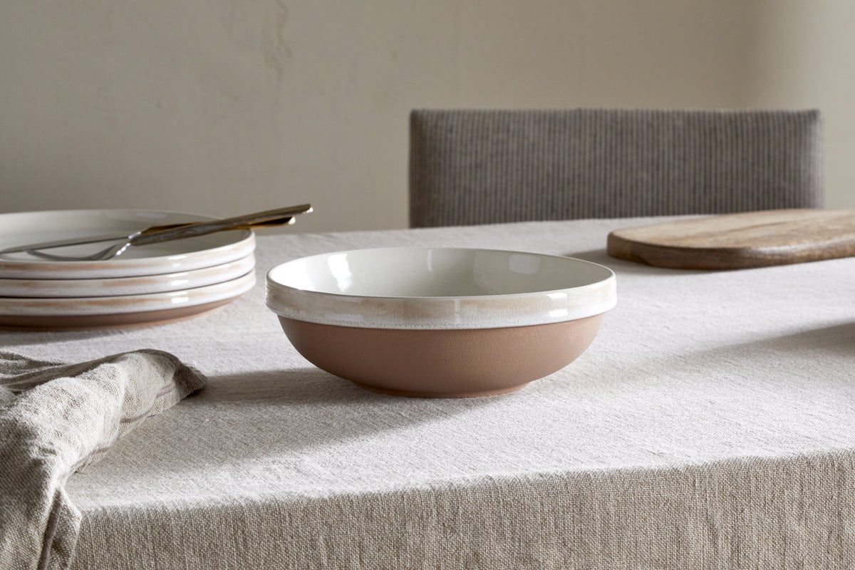 A handcrafted nkuku Edo Pasta Bowl - Terracotta with a white interior and terracotta exterior rests on a light linen tablecloth, surrounded by minimalist tableware—stacked plates, fork, wooden board—and a gray upholstered chair in a simple setting.