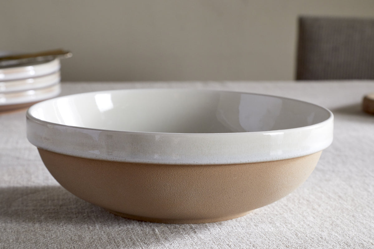 The Edo Pasta Bowl - Terracotta by nkuku, featuring a matte light brown base and glossy white rim, rests on a beige tablecloth. Minimalist tableware and a cushioned chair in the background create a calm, serene setting.