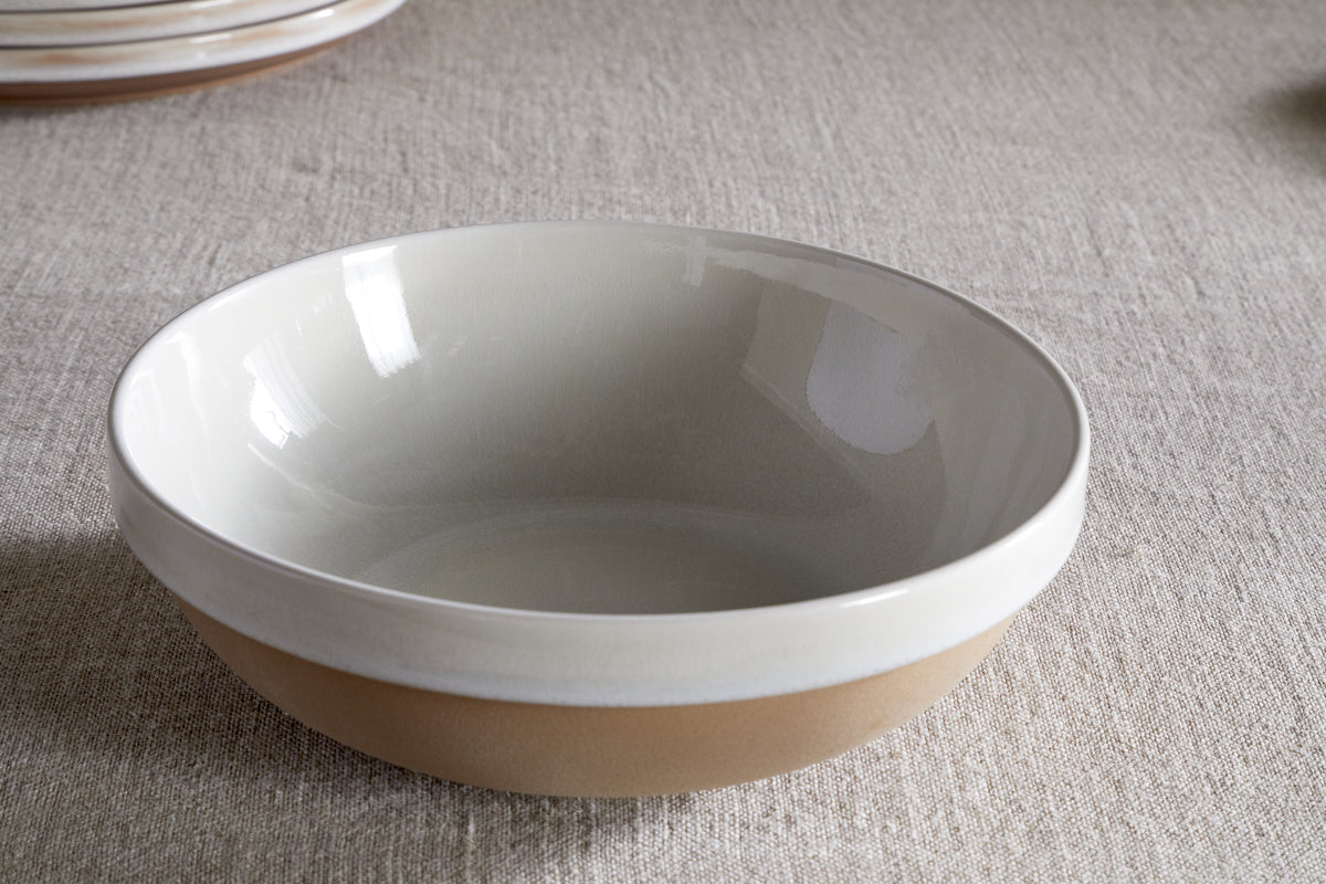 The nkuku Edo Pasta Bowl - Terracotta, featuring a handcrafted round shape with a glossy white interior and matte beige exterior, sits empty and centered on textured light beige fabric, illuminated by soft natural light.