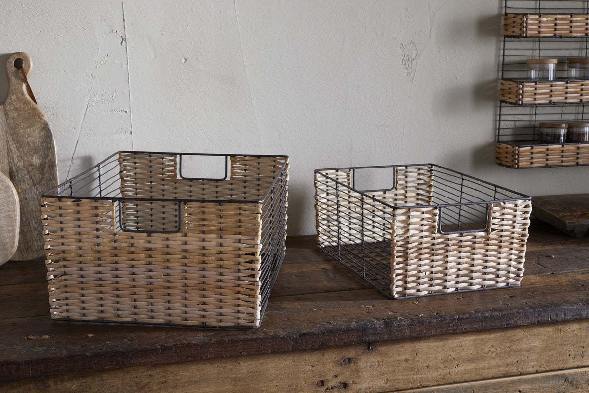 Two Montali Iron & Rattan Storage Baskets by nkuku, one larger than the other, sit on a rustic wooden surface. In the background are a wooden cutting board and shelves with small jars against a textured wall.
