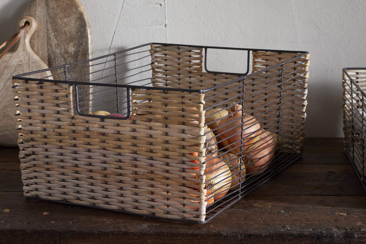 The nkuku Montali Iron & Rattan Storage Basket in natural sits on wood, filled with yellow and brown onions. A wooden cutting board rests against a white wall behind the handwoven basket.
