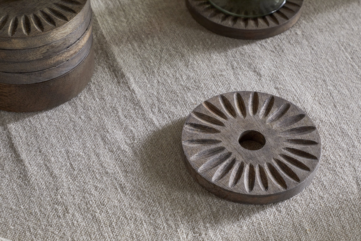The Eisha Mango Wood Coasters by nkuku, in dark brown with hand-carved sunburst design, rest on textured beige fabric. A stacked set of six and a partially visible coaster share the elegant detailing.