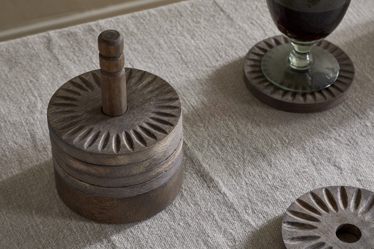 A round, carved wooden container with a handle rests on a beige textured tablecloth. Next to it is the nkuku Eisha Mango Wood Coasters - Dark Brown (Set of 6), featuring sunburst patterns and hand-carved edging; one holds a glass of dark liquid.