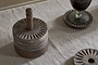 A round, carved wooden container with a handle rests on a beige textured tablecloth. Next to it is the nkuku Eisha Mango Wood Coasters - Dark Brown (Set of 6), featuring sunburst patterns and hand-carved edging; one holds a glass of dark liquid.