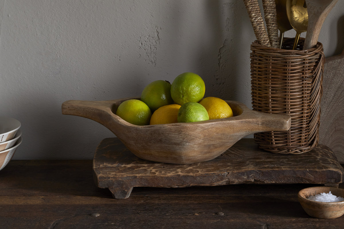 The Naashta Handled Serving Bowl - Natural by nkuku, filled with green limes and yellow lemons, sits on a rustic board, flanked by wicker utensils in a basket and stacked dishes—perfectly arranged for a cozy kitchen display.