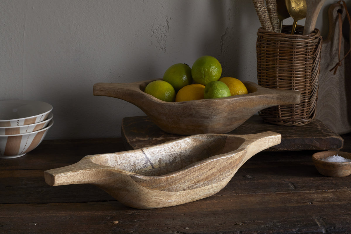 A rustic wooden dining table features two nkuku Naashta Handled Serving Bowls - Natural, one brimming with green and yellow citrus. A wicker utensil holder and stacked bowls in the background complete this cozy, earthy kitchen scene.