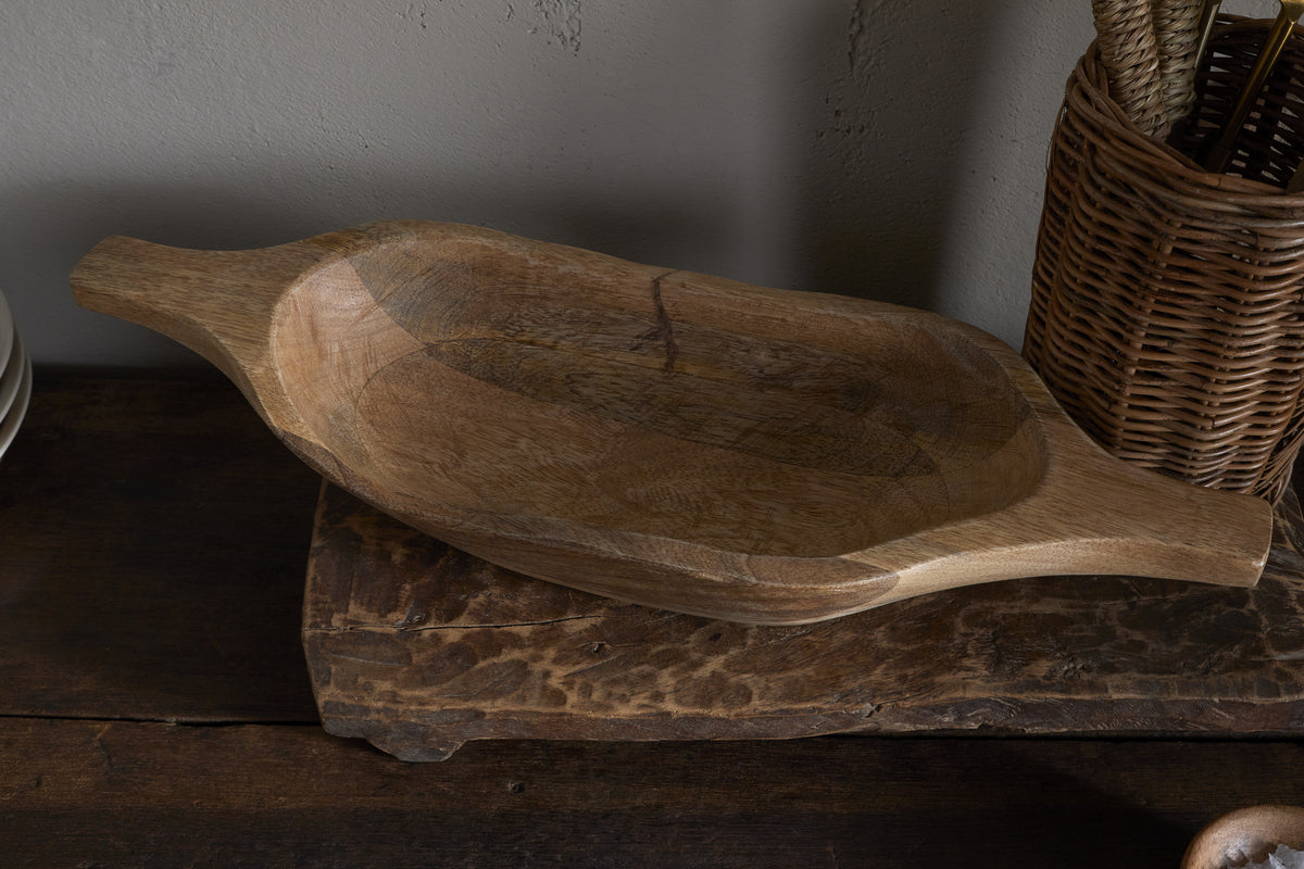 The Naashta Handled Serving Bowl - Natural by nkuku, an oval-shaped, hand-carved wooden bowl with handles, rests empty on a rustic kitchen surface. A woven utensil basket stands nearby against a neutral textured wall.