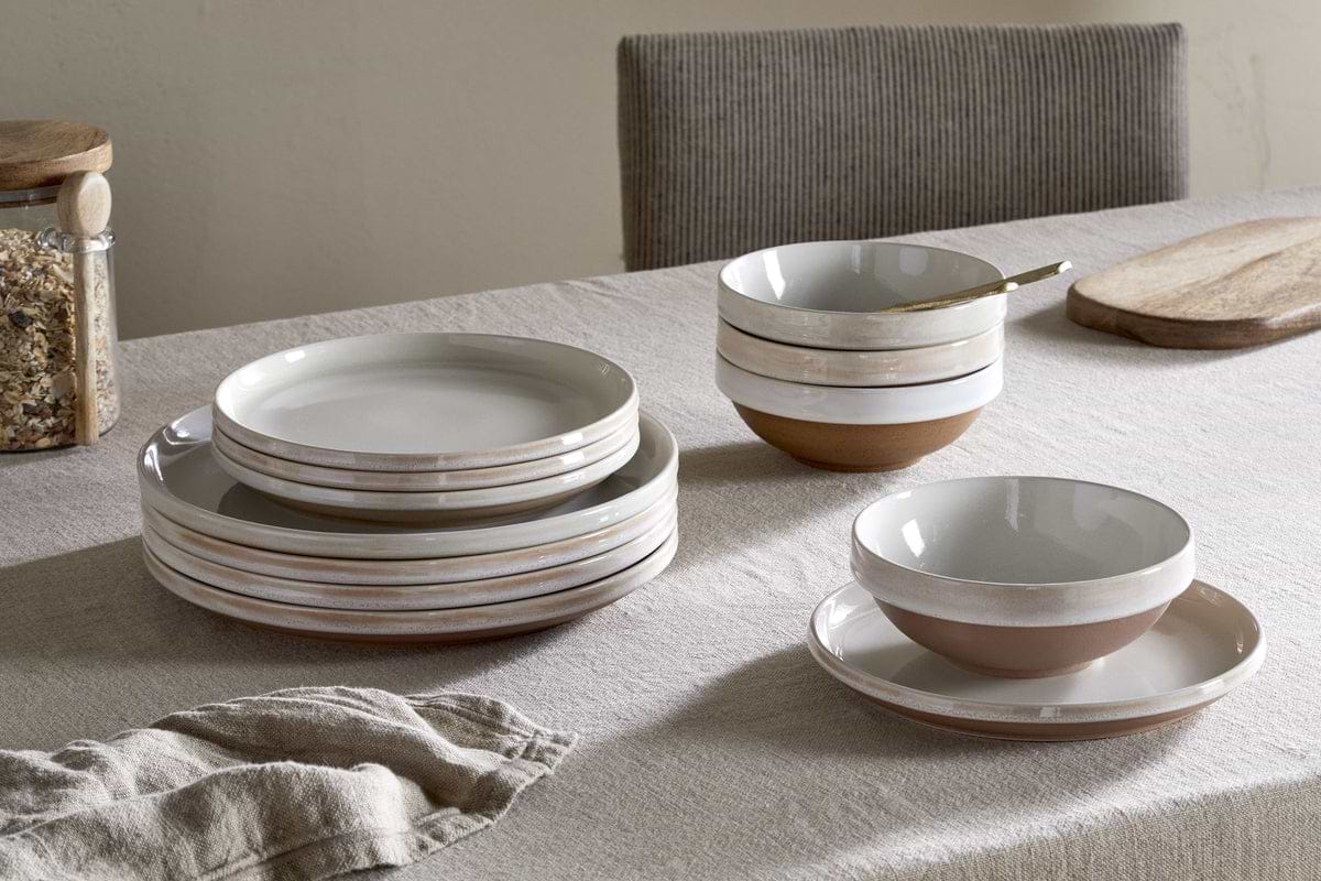 A neutral-toned table features the nkuku Edo Dinnerware Set in terracotta—stacked ceramic plates, bowls, and cups. A wooden cutting board, jar of grains, and folded napkin complete the rustic arrangement.