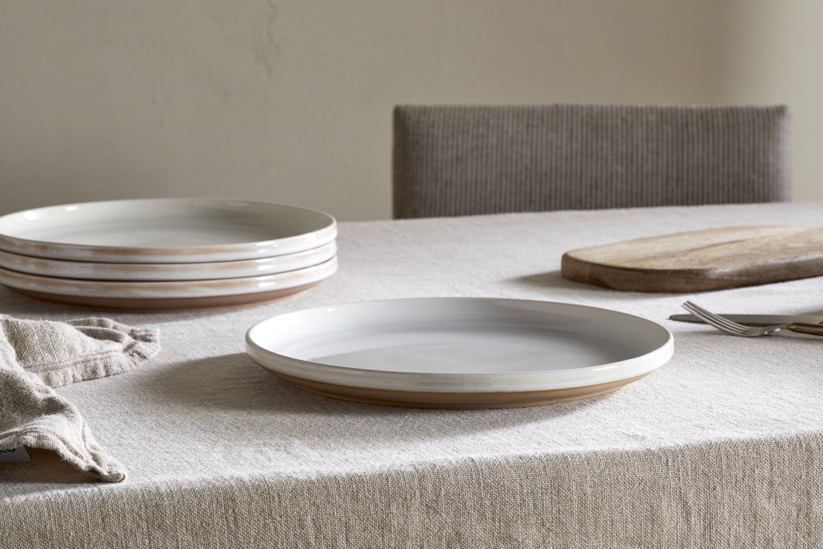 A neatly set table with a beige tablecloth showcases the nkuku Edo Dinnerware Set - Terracotta (set of 12), featuring minimalist ceramic plates, a wooden cutting board, a folded napkin, cutlery, and a cushioned chair in the background.