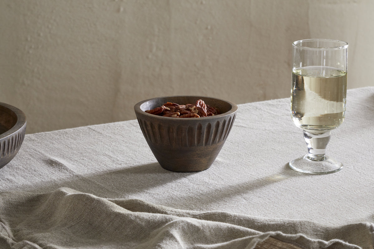 The nkuku Eisha Serving Bowl – Brown, filled with pecans, rests on a light tablecloth beside a stemmed glass of white wine and a beige napkin, creating an elegant display against a simple textured wall.