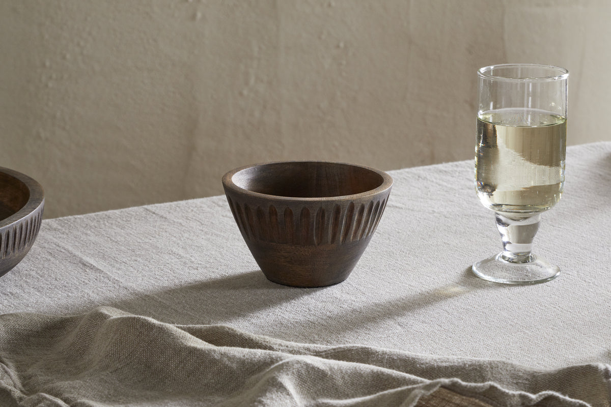 The nkuku Eisha Serving Bowl - Brown, featuring carved vertical grooves, sits on a beige tablecloth next to a glass of white wine. Soft light and faint shadows enhance the rustic, minimalist scene with another Eisha bowl partly visible on the left.