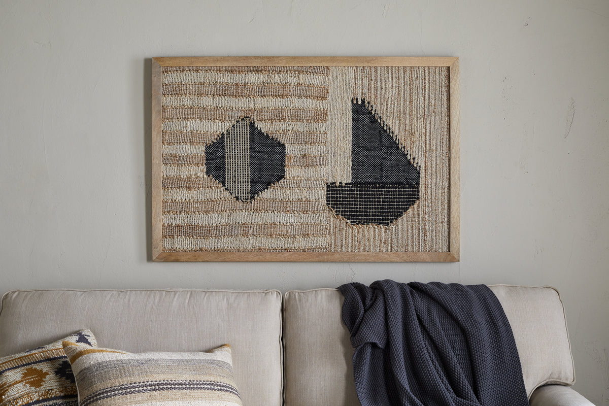 The Kiran Large Textured Wall Art by nkuku, featuring abstract dark blue geometric shapes on natural and cream stripes, hangs above a light beige couch with a gray knit blanket and a decorative geometric pillow.