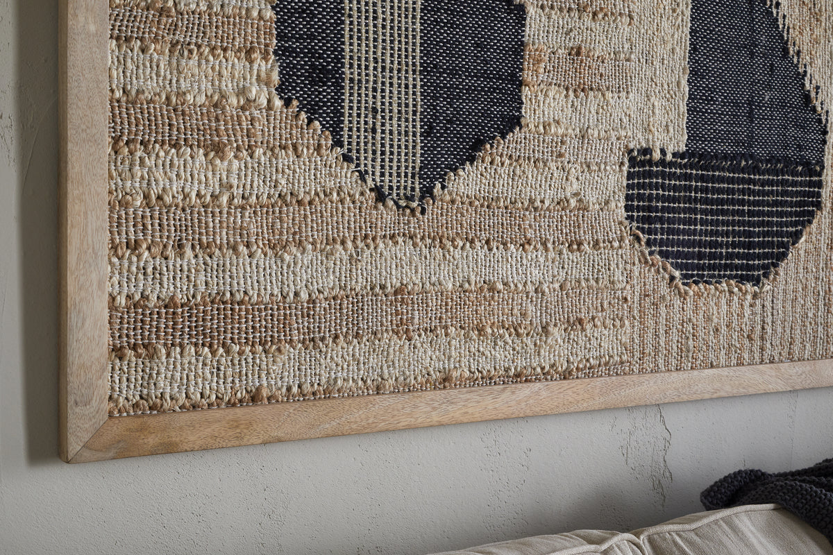The Kiran Large Textured Wall Art by nkuku features handwoven beige fibers with bold dark blue geometric shapes, framed and displayed on a light gray wall above a beige fabric sofa.