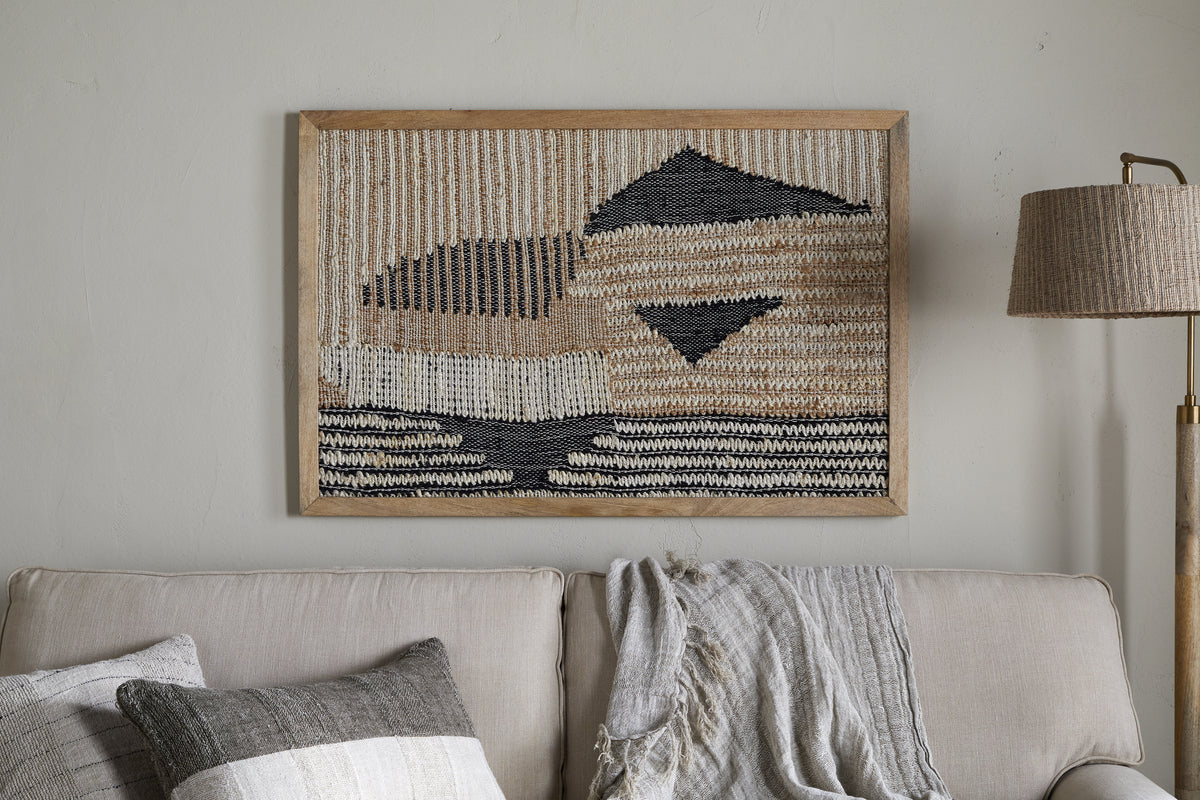 The Birala Large Textured Wall Art by nkuku, featuring natural and dark blue abstract patterns, hangs above a light beige sofa with two textured pillows and a gray throw. A floor lamp with a woven shade stands to the right.
