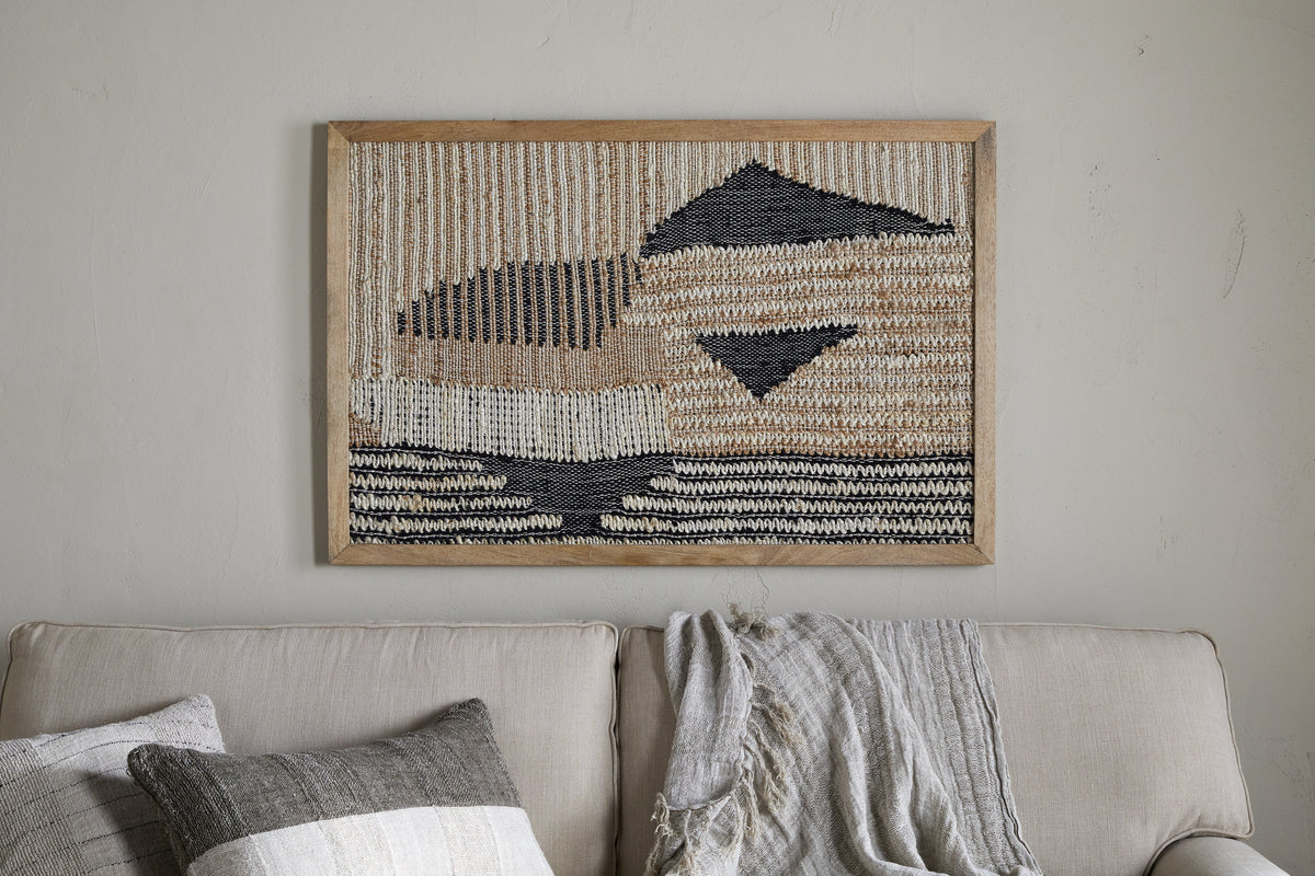 The Birala Large Textured Wall Art by nkuku, featuring natural and dark blue geometric patterns, hangs above a light beige sofa adorned with textured neutral pillows and a gray-beige throw blanket against a plain light wall.