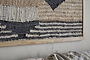 A close-up of nkuku's Birala Large Textured Wall Art in natural and dark blue hangs on a light wall. Black, cream, and brown abstract geometric patterns stand out. Below, a beige textile is draped over the couch.
