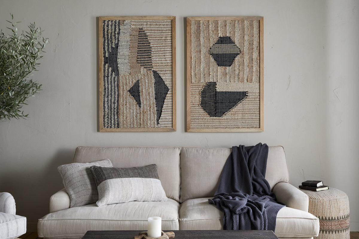 A beige sofa with neutral cushions and a dark throw sits against a light wall. Above, two Birala Large Textured Wall Art pieces by nkuku in natural and dark blue hang side by side. A side table and stacked books complete the cozy decor.