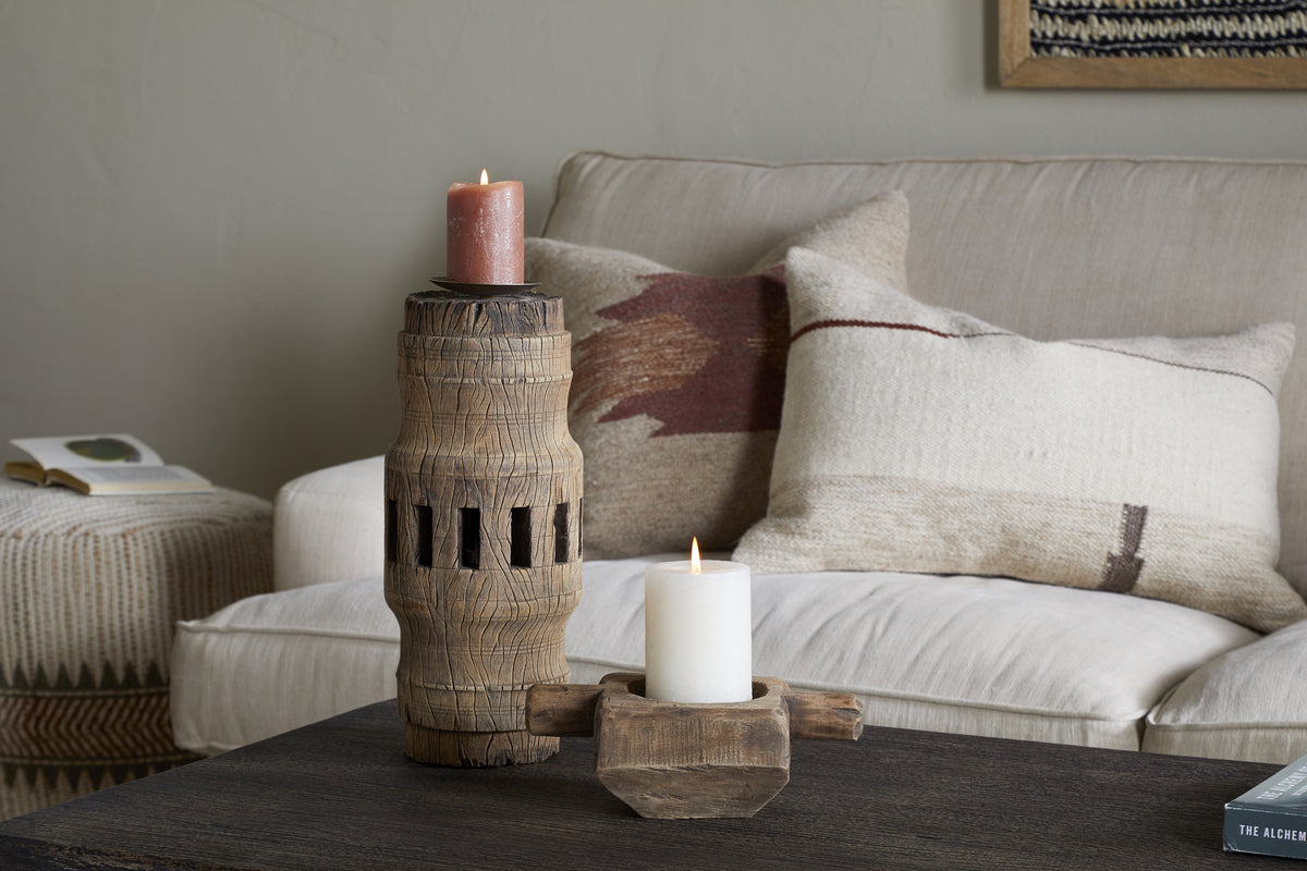 A cozy living room features a beige sofa with textured pillows. On a dark wood coffee table sit two nkuku Mvua Reclaimed Wood Pillar Candle Holders—one tall with a red candle, the other short with a white—plus books on nearby side tables.