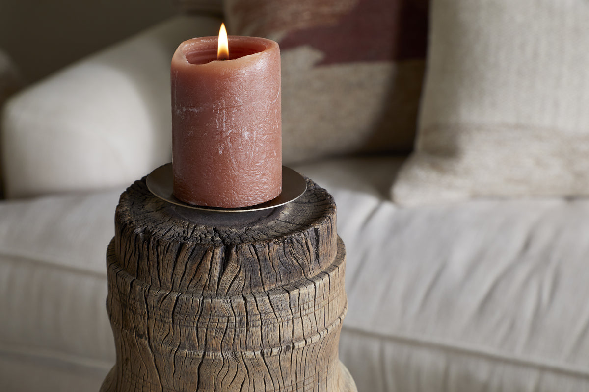 A pinkish-brown pillar candle sits on the nkuku Mvua Reclaimed Wood Pillar Candle Holder - Natural, featuring a metal plate and rustic wooden base. A white couch with neutral, textured pillows adds to the cozy ambiance in the blurred background.