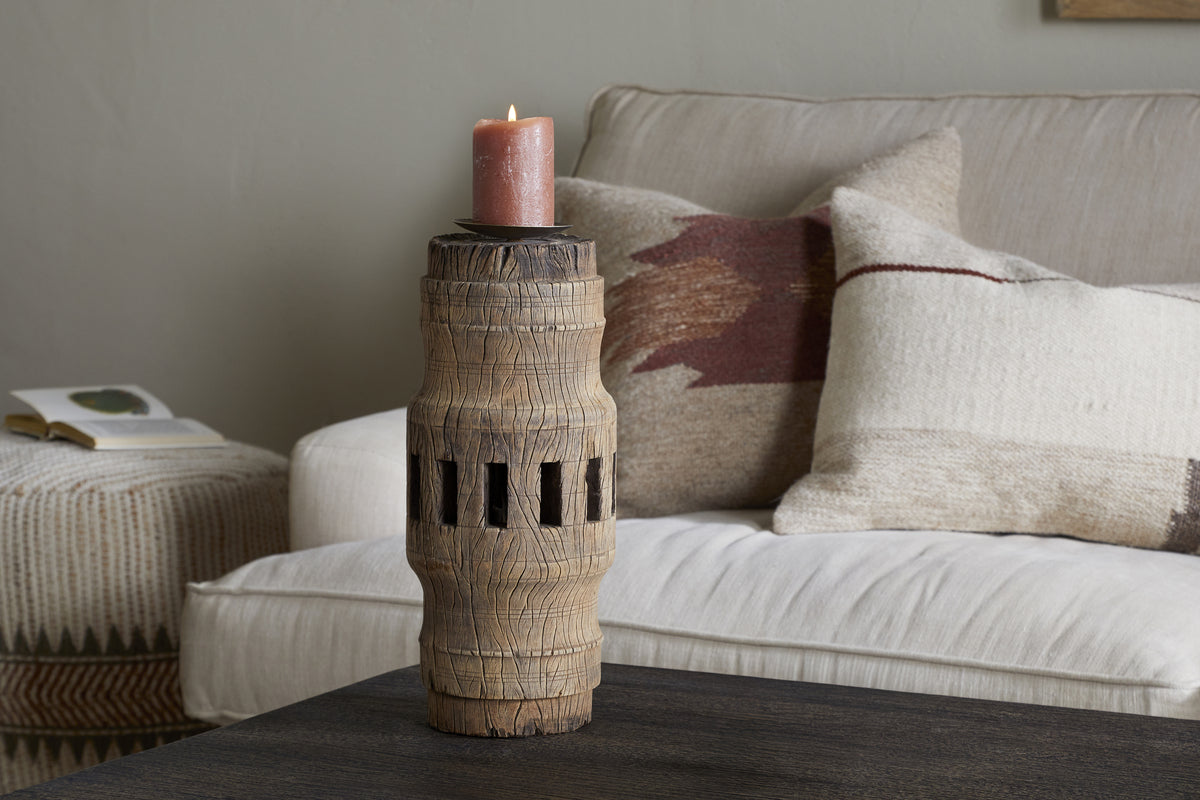 The Mvua Reclaimed Wood Pillar Candle Holder - Natural by nkuku, featuring carved grooves, sits on a dark coffee table with a lit pillar candle. A beige sofa and patterned pillows in the background add to the cozy atmosphere.