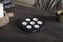 The nkuku Maji Stone Multi Tealight Holder in Antique Black with seven white candles sits on a dark wooden table beside a green ceramic mug, a large white pillar candle on a wooden tray, and a gray textured blanket over a beige couch.