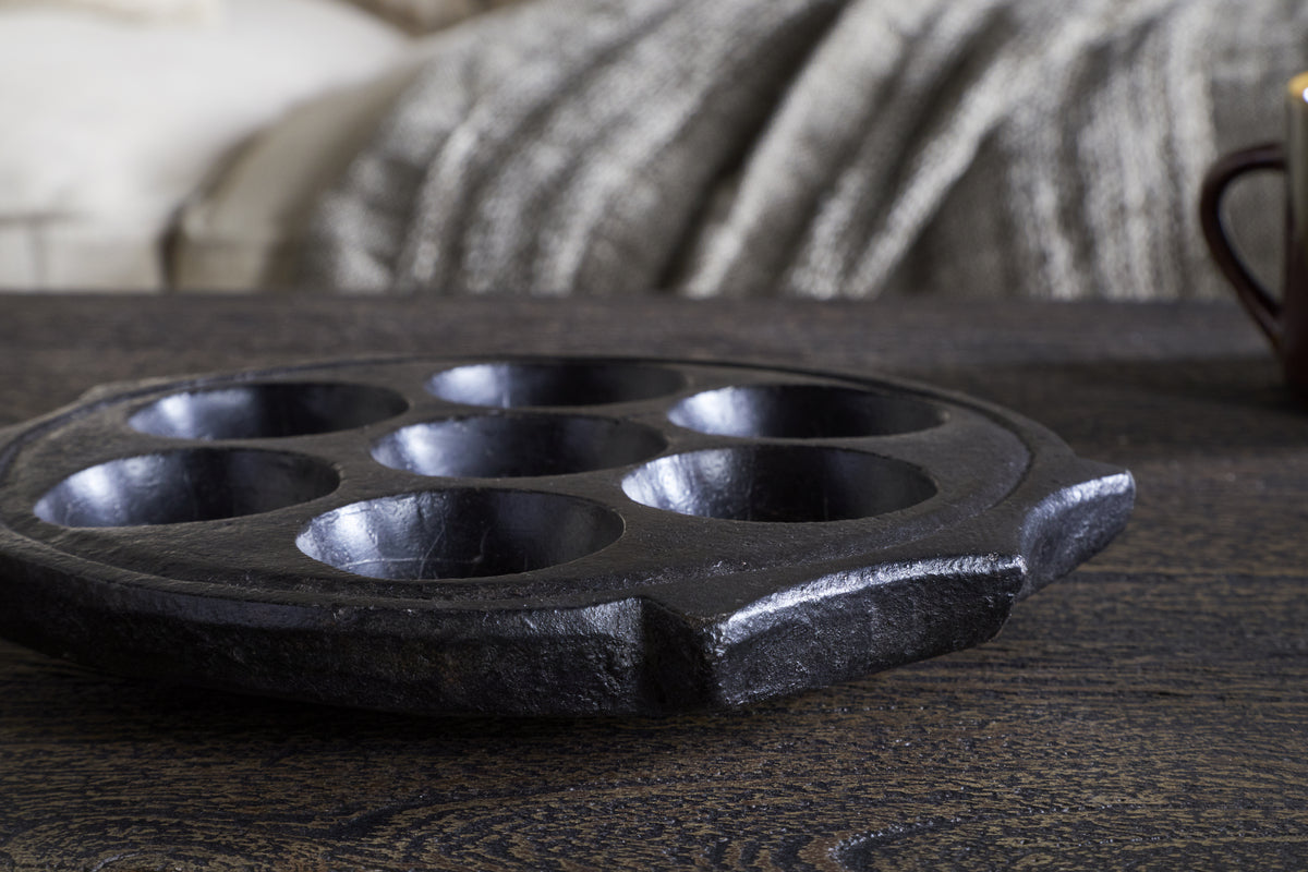 A close-up of the Maji Stone Multi Tealight Holder - Antique Black by nkuku sits on a dark wooden table. In the background are a textured gray blanket, cream sofa, and a partially visible brown mug to the right.