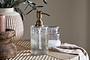 The Ilcoso Recycled Hammered Glass Soap Dispenser - Clear by nkuku features an antique brass pump and sits on textured wood, accompanied by a glass jar, striped cloth, ribbed ceramic vase with foliage, and a brown candle holder for a rustic look.