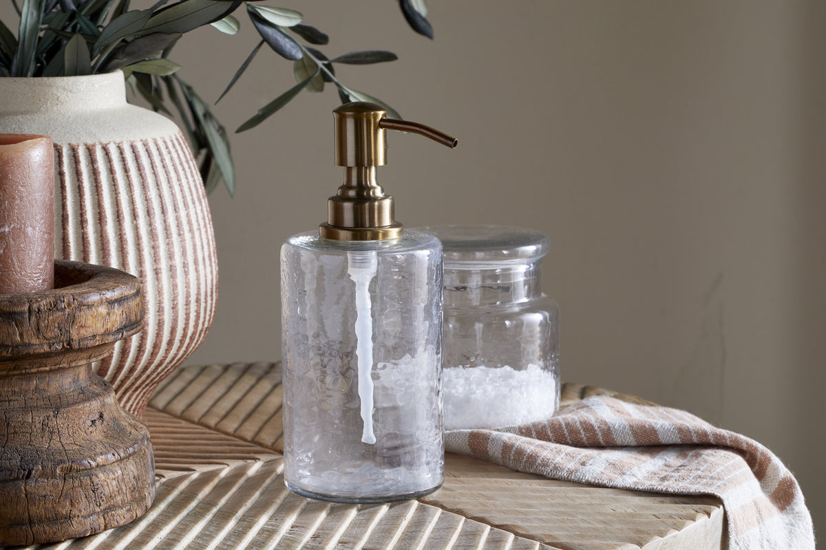 The Ilcoso Recycled Hammered Glass Soap Dispenser by nkuku, featuring an antique brass pump, rests on a textured beige surface beside a salt jar, striped towel, wooden stand with candle, and cream vase with greenery for a cozy, natural feel.