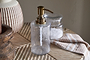 The nkuku Ilcoso Recycled Hammered Glass Soap Dispenser in clear, featuring an antique brass pump, rests on a wooden surface beside bath salts and a tan and white striped towel, with a textured vase and wooden decor in the background.