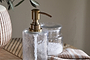 The Ilcoso Recycled Hammered Glass Soap Dispenser in clear by nkuku, featuring an antique brass pump, brings elegant style and sustainability to your bathroom.