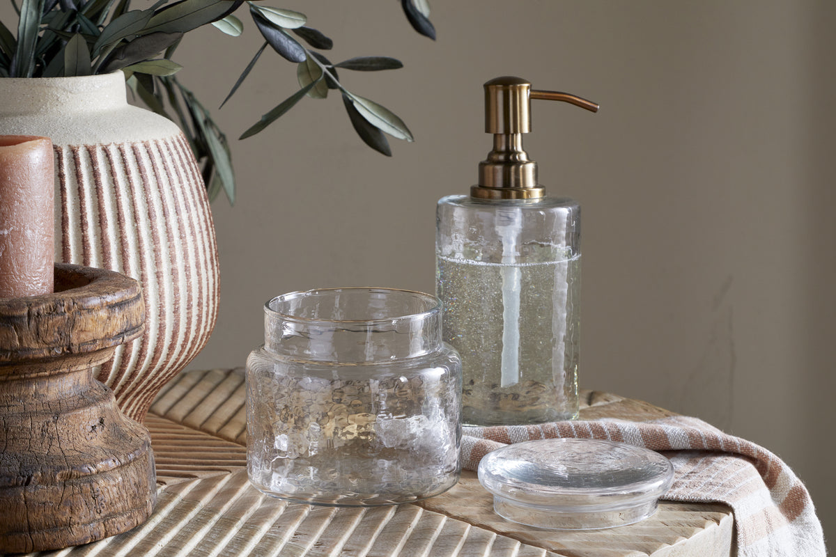 The nkuku Ilcoso Recycled Hammered Glass Storage Pot in clear sits open on wood beside a glass soap dispenser with brass pump, striped vase of green leaves, and rustic candle holder—ideal for stylish bathroom storage in natural light.