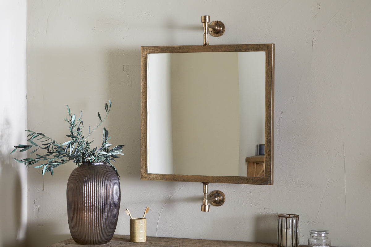 The nkuku Visala Swivel Wall Mirror in Antique Brass is mounted on a beige wall above a wooden table holding a ribbed brown vase with green branches, a gold brush cup, glass container, and metallic candle holder.