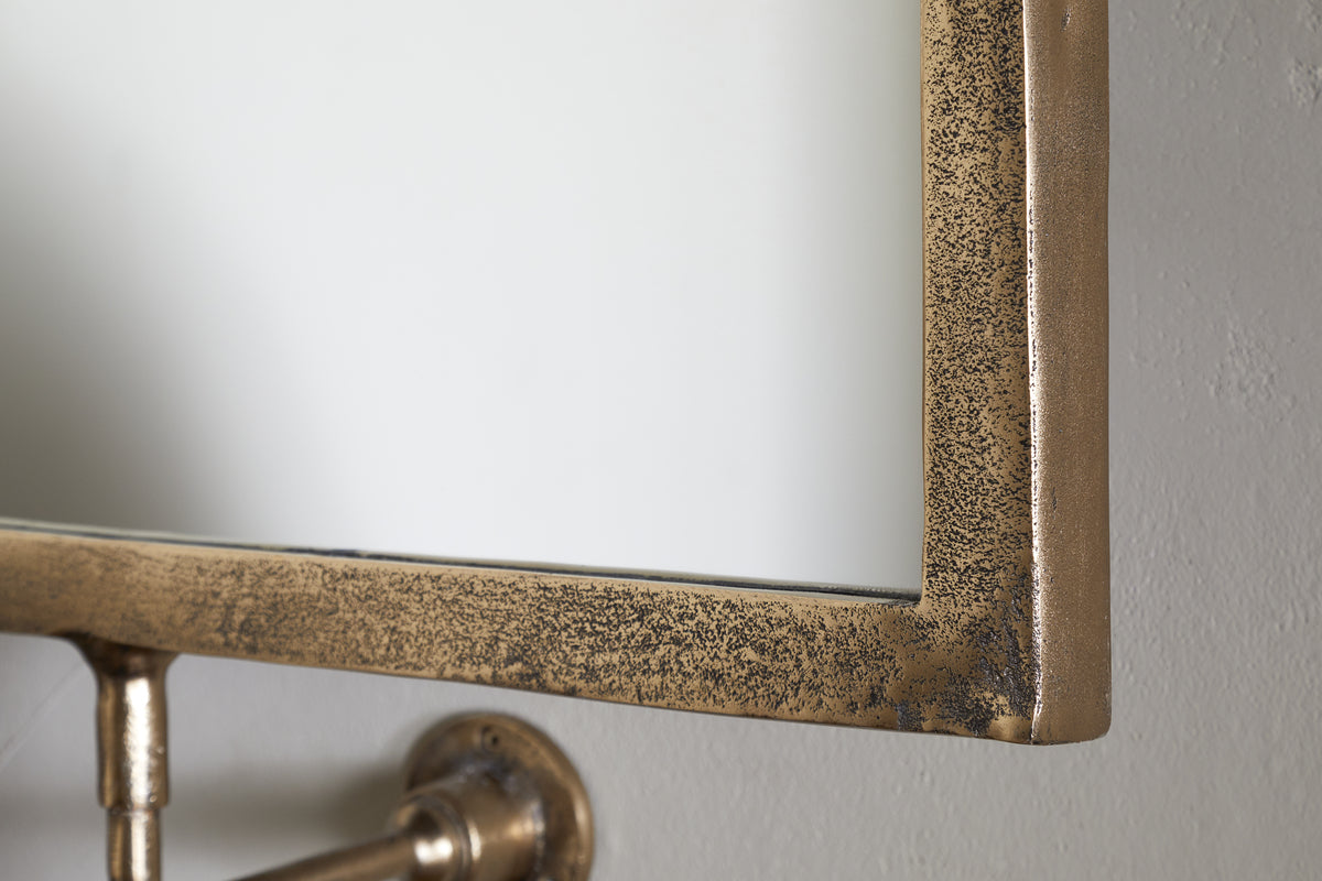 Close-up of the Visala Swivel Wall Mirror by nkuku, featuring a thick, textured antique brass frame. The mirror is mounted on a light gray wall with a metal bracket, reflecting mostly white for a rustic accent.