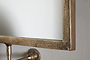 Close-up of the Visala Swivel Wall Mirror by nkuku, featuring a thick, textured antique brass frame. The mirror is mounted on a light gray wall with a metal bracket, reflecting mostly white for a rustic accent.